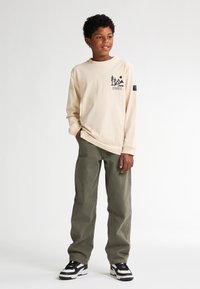 Tricou bej cu mânecă lungă, cu imprimeu grafic, purtat cu pantaloni verzi măsliniu și adidași negri și albi. Croială simplă, lejeră.