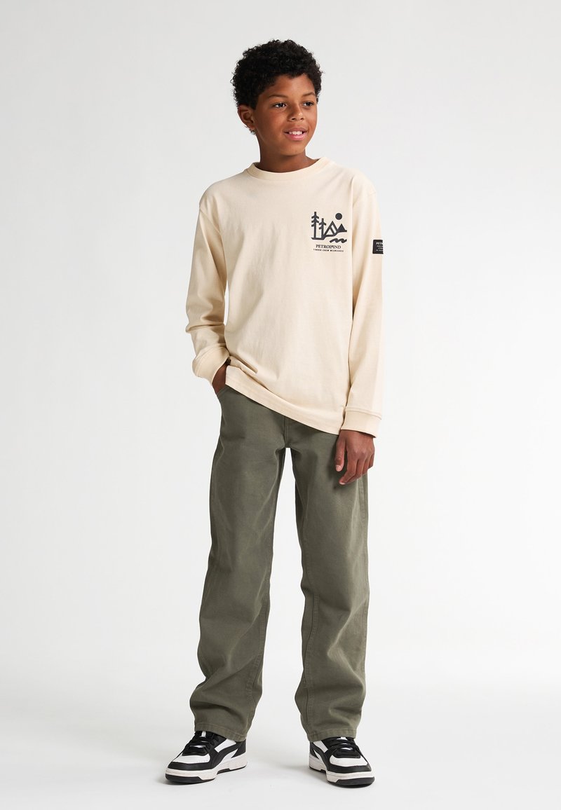 Tricou bej cu mânecă lungă, cu imprimeu grafic, purtat cu pantaloni verzi măsliniu și adidași negri și albi. Croială simplă, lejeră.