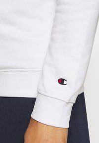 Poignet de sweat-shirt à manches longues blanc avec un petit logo Champion noir et rouge au-dessus du poignet sur le bras d'une personne.
