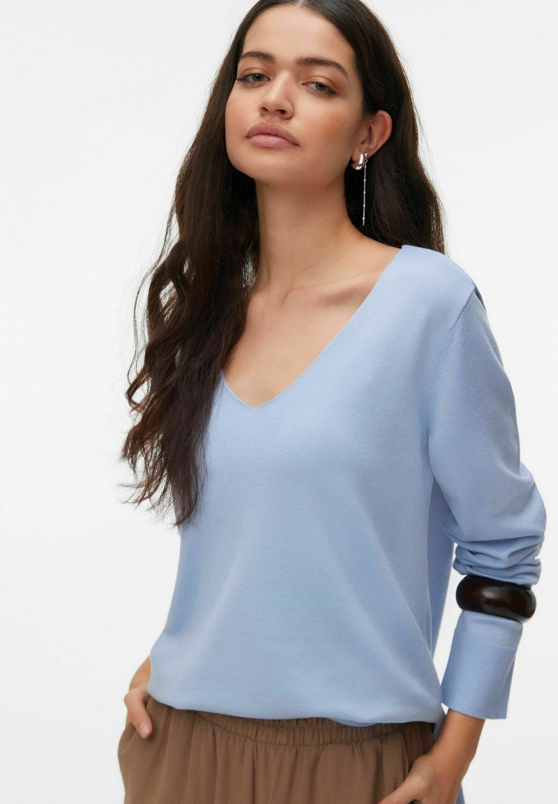 Pull léger bleu clair à col en V avec manches longues, texture douce et poignets ajustés. Associé à une jupe à taille élastique marron, présentant un design simple.