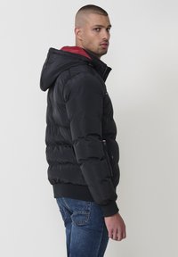 Svart puffjacka med huva som har röd kant, med dragkedja fram och elastiska muddarna. Bärs med blå jeans, visad från en sida.