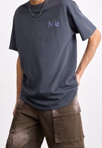 Mörkgrå bomullströja med en lila Nike-logotyp på vänster bröst, tillsammans med bruna cargoshorts. Strukturerad tyg, avslappnad passform.