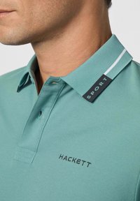 Muž v tyrkysovém polo triku s vyšitým nápisem "HACKETT" na hrudi a štítkem "SPORT" na límci.