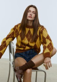 Brun og gul argyle strikket sweater med ribbet halsudskæring og manchetter, parret med denimshorts og brune ankelstøvler på en metalstol.