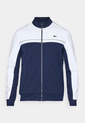 Marineblauw en wit hesje met rits, hoge kraag, lange mouwen en een klein groen krokodillenlogo op de borst.