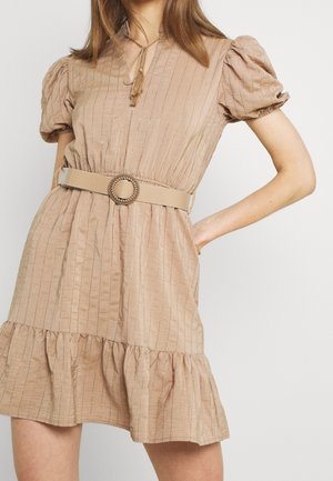 Vrouw die een beige jurk met korte mouwen draagt, met opgerolde mouwen, een V-hals, een geplooide taille, een ronde gesp in de taille, en een gerimpelde zoom.