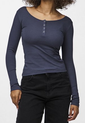 Kvinde iført mørkeblå ribstrikket henley-bluse med lange ærmer og tre knapper samt sorte højtaljede jeans, stående foran en ensartet baggrund.