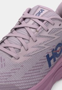 Αθλητικό παπούτσι σε λιλά χρώμα με υφασμάτινη υφή, στρατηγικά στερεωμένα κορδόνια σε σχήμα σταυρού και λογότυπο "HOKA" στο πλάι, που αναδεικνύει τις λεπτομέρειες σχεδίου στην κορυφή και στο πλάι.