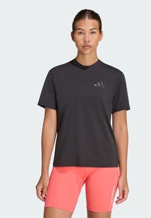 Kobieta ubrana w ciemnoszarą koszulkę Adidas i jaskrawo koralowe krótkie spodenki sportowe stoi na tle jednolitego, jasnego tła.