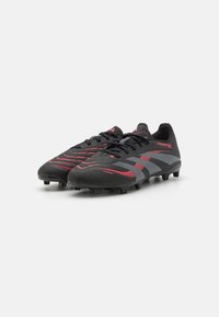 Botas de fútbol negras con acentos rojos y grises, que cuentan con una parte superior texturizada, cordones y tacos moldeados para tracción en el campo.