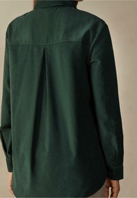 Camicia verde scuro a maniche lunghe con pince sul retro, colletto e texture liscia. Vestibilità sartoriale con polsini abbottonati.
