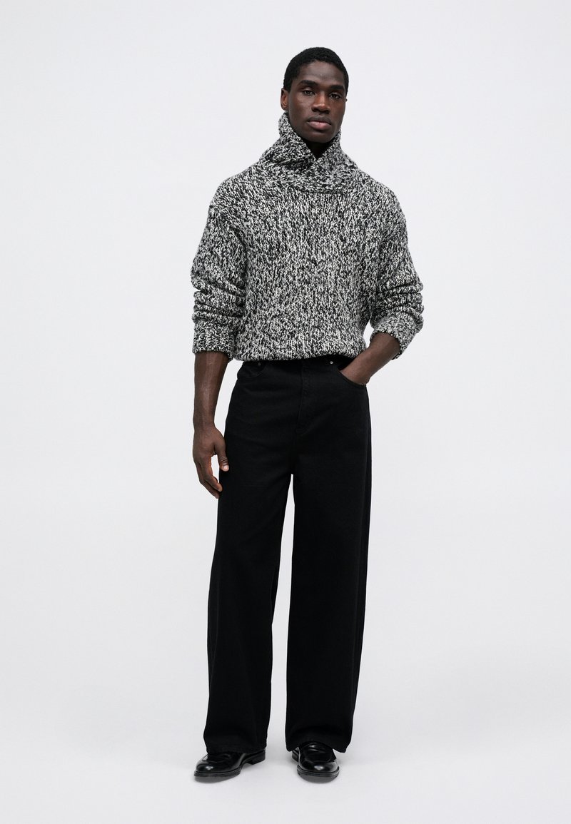 Maglione lavorato a maglia spesso in bianco e nero, con colletto alto e vestibilità rilassata, abbinato a pantaloni neri a gamba larga e scarpe.