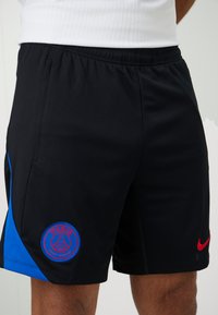 Sorte atletiske shorts med et struktureret stof, blå sidepanel, rødt Nike-logo og et rundt Paris-logo på den nederste venstre ben.