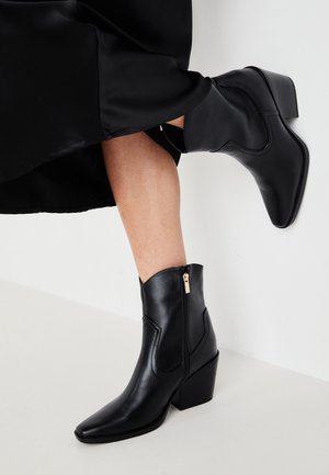 Bottes cheville en cuir noir avec fermeture éclair latérale et talon carré portées avec une jupe noire sur fond clair.