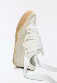 Fehér tornacipő sima anyagból, texturált oldalpanelekkel és vastag fehér fűzőkkel, gumitalpú, lekerekített orral.