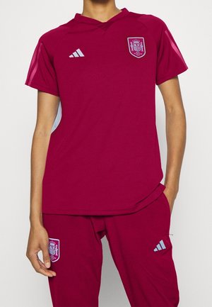 Osoba ubrana w bordowy strój piłkarski Hiszpanii, zarówno koszulkę, jak i spodnie, z widocznym logo Adidas oraz herbem narodowej drużyny na piersi i udzie.