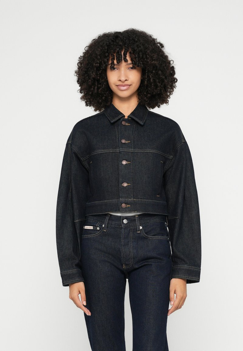 Krótka kurtka dżinsowa w ciemnym niebieskim kolorze z zapięciem na guziki, oversize'owymi rękawami i dwoma kieszeniami na piersiach. Gładka faktura i klasyczny design dżinsu.