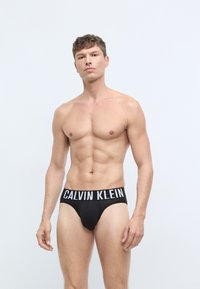 Crne muške gaćice od pamuka s širokom elastičnom gumom na kojoj je bijeli natpis "CALVIN KLEIN". Glatka tekstura, udoban kroj i klasičan dizajn.
