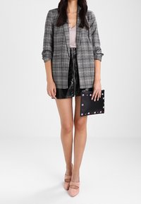 Šedé kárované sako přes světlý top, doplněné lesklými černými šortkami. Drží černé psaníčko s barevnými akcenty, na nohou má růžové lodičky.