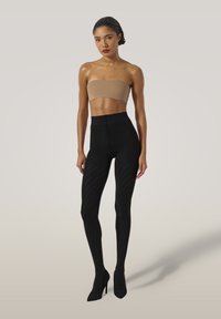 Kvinna i en beige bandeau-top, svarta mönstrade tights med hög midja och svarta högklackade skor som står mot en ljus bakgrund.