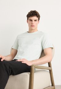 Camiseta de manga curta verde menta clara com um pequeno emblema e decote redondo, combinada com calças pretas, sentado numa cadeira.