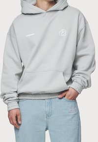 Sweat à capuche gris avec une poche kangourou, des poignets et un ourlet côtelés. Présente un petit logo sur la poitrine et un emblème rond sur le côté gauche.