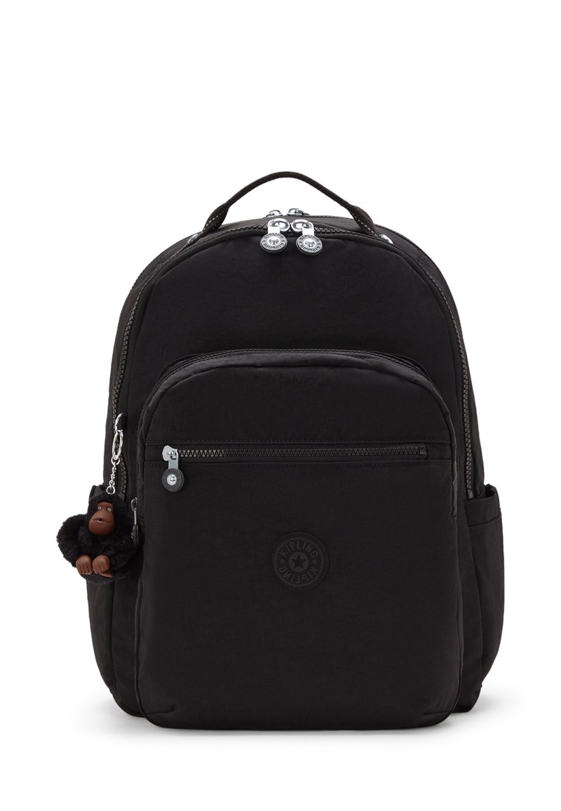 Mochila negra con tejido texturizado, cremalleras dobles, bolsillo frontal y un llavero de mono como acento. Presenta un logo circular de la marca en la parte frontal.