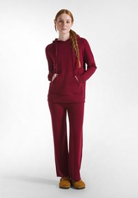 Hanorac cu glugă texturat de culoare burgundă, cu buzunar frontal, asortat cu pantaloni largi din același material; încălțăminte de culoare portocalie-maro.