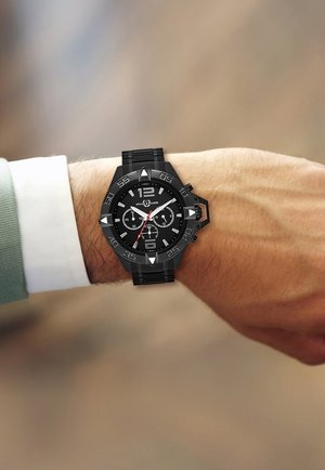 Black sports watch with a textured black dial, silver accents, three sub-dials, and a rubber strap, displayed on a wrist with a light green shirt.