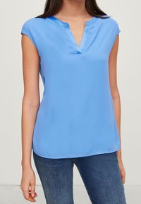 Helles blaues Kurzarmbluse mit V-Ausschnitt und glatter, leichter Textur. Kombiniert mit lässigen blauen Jeans.