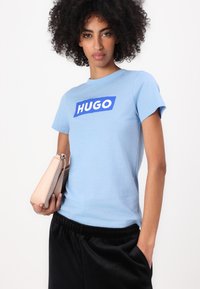 Modrá bavlnená tričko s neónovo modrým obdĺžnikovým logom a bielym textom "HUGO." V páre s čiernymi teplákovými nohavicami.
