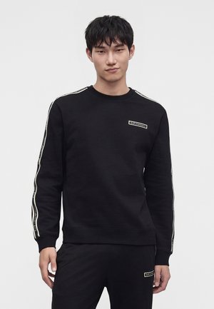 Jeune homme aux cheveux courts noirs portant un sweat-shirt noir et un pantalon avec des bandes blanches à logo sur les manches et la poitrine.