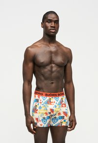 Μυώδης μαύρος άνδρας στέκεται μπροστά σε απλό φόντο φορώντας πολύχρωμα boxer briefs με σχέδια και πορτοκαλί ζώνη μέσης με λογότυπο Björn Borg.