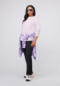 Lichtpaarse sweatshirt, zwarte broek en witte sneakers. Een geknoopte paarse geblokte shirt en oversized zonnebril maken de look compleet.