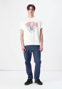 Hvid bomulds T-shirt med "TOMMY JEANS" grafik, parret med blå denimjeans og sorte sneakers, stående mod en ensfarvet baggrund.