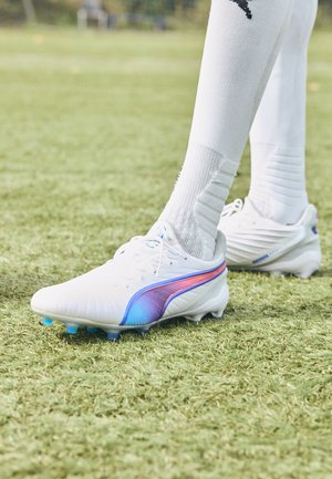 Des crampons de football blancs avec une bande dégradée violette-bleue, présentant un design épuré, un dessus texturé et des crampons bleus contrastants sur la pelouse.