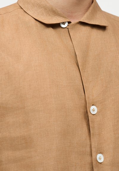Chemise en lin marron clair avec un col arrondi et des boutons blancs. Le tissu présente une texture qui souligne sa qualité respirante.