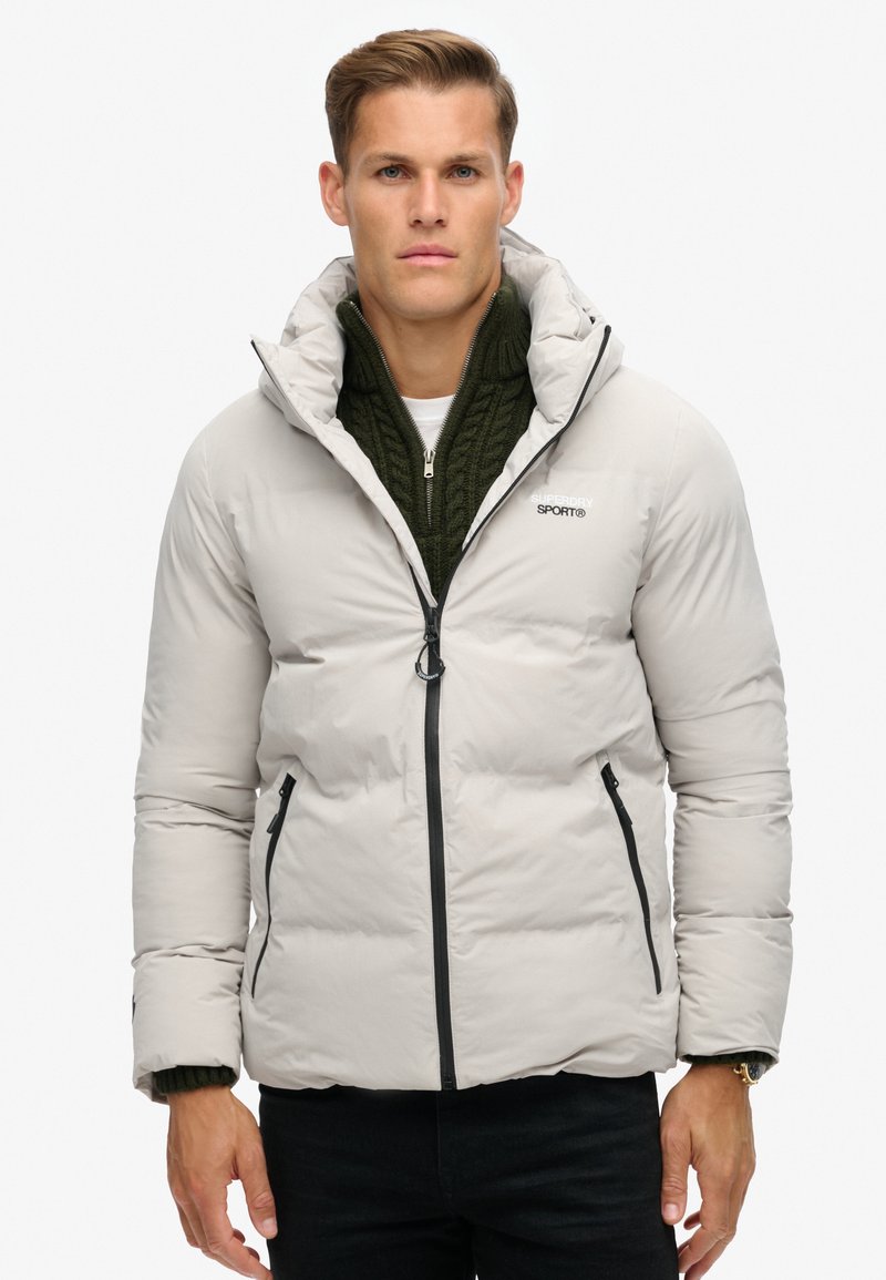 Lichtgrijze pufferjack met een hoge kraag, een rits aan de voorkant en twee zwarte ritszakken aan de zijkant. Draagt over een donkergroen gebreid sweater.