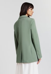 Femme aux cheveux foncés portant un blazer vert sauge vu de dos, associé à une jupe blanche, sur un fond clair et uni.