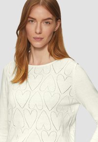 Pull tricoté blanc avec des découpes en forme de cœur dans un motif répétitif, col rond et manches longues. Texture lisse.