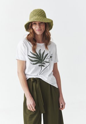 T-shirt blanc avec des imprimés de plantes vertes et de fleurs, associé à un pantalon large taille haute de couleur vert olive. Le modèle porte un chapeau texturé vert.