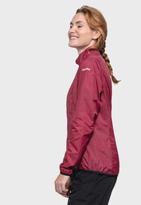 Rote leichte Jacke mit hohem Kragen, Reißverschluss vorne und elastischen Bündchen. Verfügt über ein Logo am Oberarm und hat eine glatte Textur.