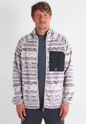 Giacca in pile color crema con motivi geometrici neri, chiusura con zip frontale e tasca sul petto nera. Maniche lunghe e texture morbida.