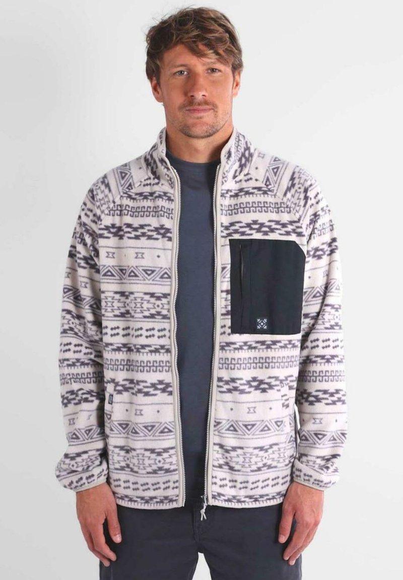 Giacca in pile color crema con motivi geometrici neri, chiusura con zip frontale e tasca sul petto nera. Maniche lunghe e texture morbida.