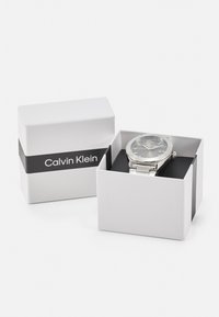 Reloj de acero inoxidable plateado con una esfera redonda y una esfera gris, presentado en una caja blanca con una correa negra que lleva "Calvin Klein".
