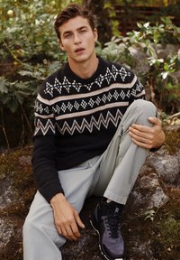 Maglione nero con motivo fair isle bianco e marrone, pantaloni grigi chiari e scarpe da trekking nere. Modello seduto su una superficie rocciosa con vegetazione.