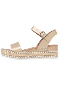 Beige Wedgesandale mit Riemen und Schnalle am Knöchel, dekorierte Plateausohle mit metallischen Nieten und geflochtenen Details.