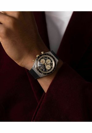 Montre chronographe avec un cadran noir, des sous-cadrans beiges, un boîtier en acier inoxydable, des accents dorés, et un bracelet en caoutchouc noir, portée au poignet.