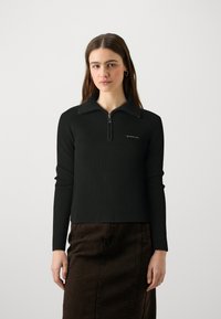 Haut noir à col zippé quart avec des accents d'épaule, présentant une petite étiquette logo sur la gauche. Associé à une jupe en denim marron.