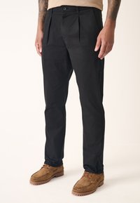 Black trousers with pleats, made of smooth fabric, feature a classic cut and a single button closure at the waist. Worn with brown suede shoes.